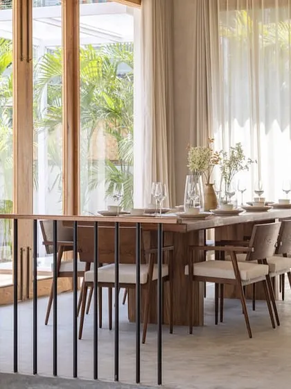 The dining area in the "House of Basho" is positioned next to large, openable wooden doors that lead out to the garden. Sheer curtains diffuse the light, creating a soft and airy atmosphere that connects the indoor mealtime experience with the tropical outdoors.