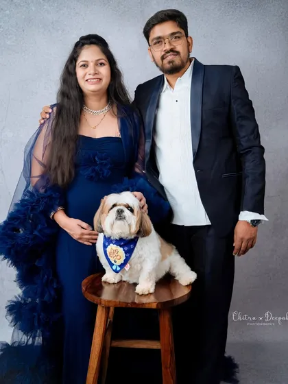 An elegant studio portrait of an expecting couple dressed in formal blue attire, with their well-behaved dog posing perfectly on a stool.