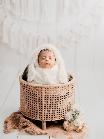 A beautiful boho-chic newborn portrait. The baby, wearing a furry bonnet, sleeps in a rattan basket against a clean, white backdrop with macrame details.