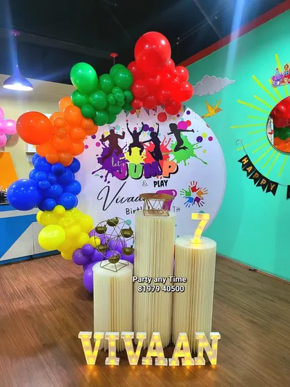 A colorful "Jump and Play" theme for Vivaan's 7th birthday. The setup includes a vibrant custom backdrop, a multi-colored balloon garland, and light-up name and number props.