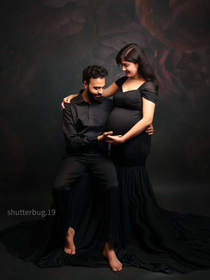 A tender couple's portrait in coordinated black outfits. The mom-to-be wears a fitted black mermaid-style gown that beautifully highlights her baby bump, creating a classic and elegant look.