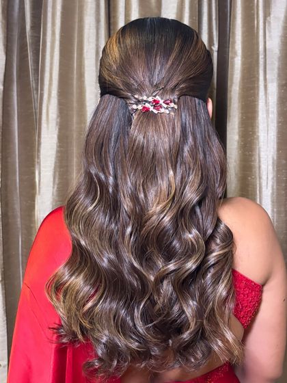 This half-up style features a simple twist secured with an elegant red and crystal clip, complementing the client's red saree. The hair has beautiful highlights and is styled in glossy, defined waves.