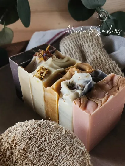 This image captures the art of slow beauty. It features a selection of my handcrafted cold-process soaps next to a natural loofah and a soap saver pouch, highlighting the traditional methods and natural accessories I offer.
