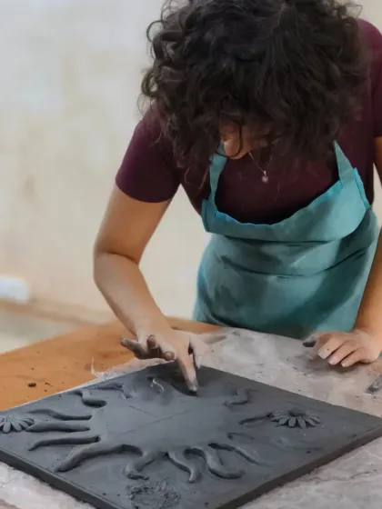 A student works on a large clay mural, sculpting a sun design. This type of project allows for exploration of texture, pattern, and large-scale composition in clay.