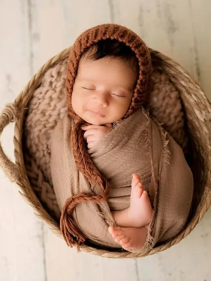 This swaddling style leaves the baby's feet peeking out, which is an adorable detail to capture. The earthy brown tones of the wrap, bonnet, and basket create a warm, organic feel.