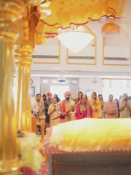 The couple stands before the Guru Granth Sahib during their Sikh wedding. This wide shot captures the entire scene, with family looking on in support.