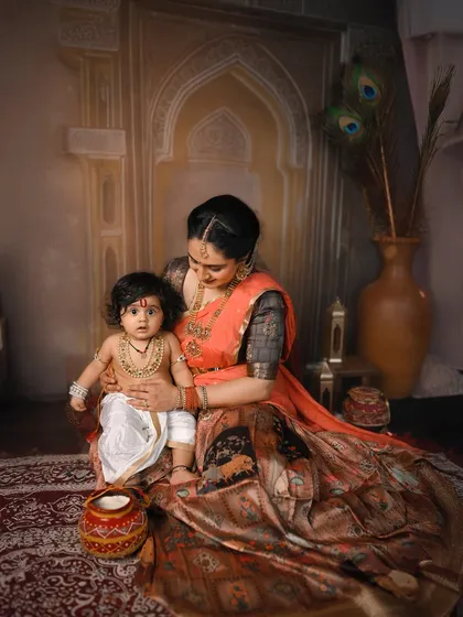 A mother looks down lovingly at her baby, dressed as little Krishna. The entire scene is set up to look like a classic painting.