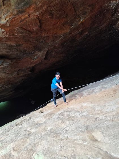 A trekker using a rope to descend a steep, smooth rock surface near a cave.