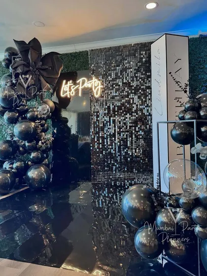 For a modern and glamorous corporate party, we designed this chic photo booth area. The setup features a black shimmer wall, a custom 'Let's Party' neon sign, and a sophisticated black balloon arrangement, perfect for milestone celebrations.