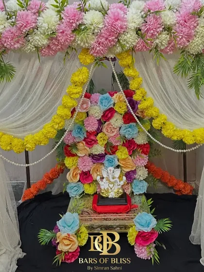 A vibrant and colorful floral backdrop for Ganesh Chaturthi. This unique design features a wall of multi-colored flowers behind the idol, framed by pink and white floral arrangements and yellow marigold garlands.