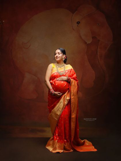 A full-length portrait of a mother-to-be in a red and gold saree, cradling her bump. The elephant backdrop adds a touch of traditional grandeur.