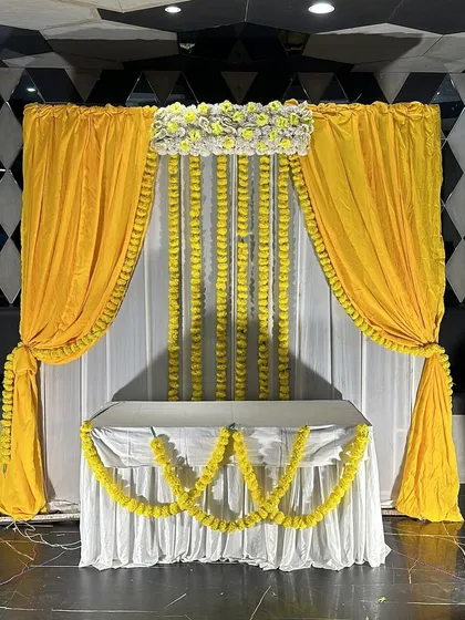 A clear, straight-on shot of the backdrop we created for a home kirtan, featuring yellow drapes, marigold garlands, and a floral header. This is a perfect example of our simple yet elegant decor packages.
