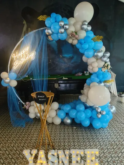 This stylish blue, white, and silver balloon arrangement is perfect for a sophisticated birthday celebration. The setup includes a circular frame with a neon 'Happy Birthday' sign and a light-up name display for 'YASNEE'.