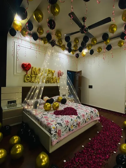 An intimate birthday room surprise shown in evening light. The combination of gold and black balloons, a fairy light canopy, and a rose petal path creates a warm and celebratory atmosphere.