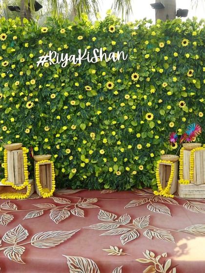 A custom photo wall for the Haldi ceremony. The lush green backdrop is dotted with sunflowers and features the couple's hashtag, creating a personalized and vibrant spot for pictures. The pink benches add a fun pop of color.