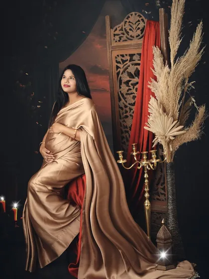 A portrait of pure elegance. This mom-to-be looks regal and serene, seated in a golden satin saree against a rich, traditionally styled backdrop.
