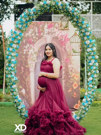 A stunning solo portrait of the mother-to-be in a maroon gown, framed by a beautiful floral arch.