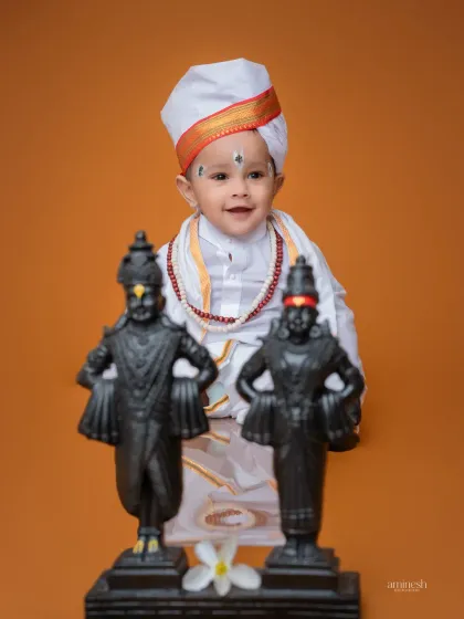 A close-up portrait of a baby boy in a Varkari costume, smiling behind idols of Vitthal and Rukmini. The simple orange background puts the focus on his adorable expression and traditional attire.