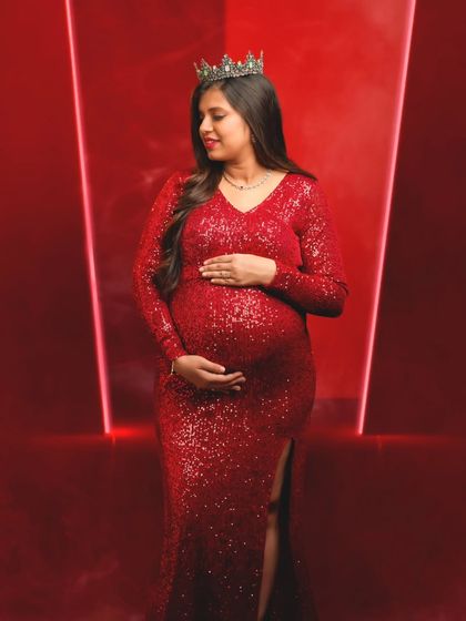 A regal and powerful portrait of a mom-to-be in a red sequin gown and crown, looking down at her bump with love.