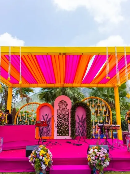Welcome to Rangeela Rajasthan! We love creating immersive themes, and for this Mehendi, we transformed an outdoor space into a vibrant carnival with bright pink and yellow drapes, traditional Rajasthani decor, and a stage ready for folk music and dancing.