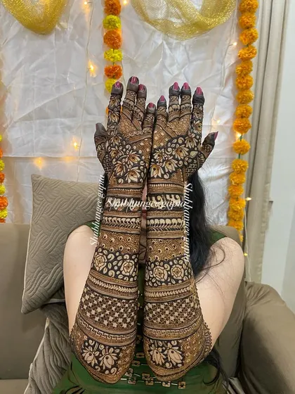 A lovely photo of a bride showcasing her full back-of-hand mehndi against a festive background. The design is filled with intricate floral and checkerboard patterns.