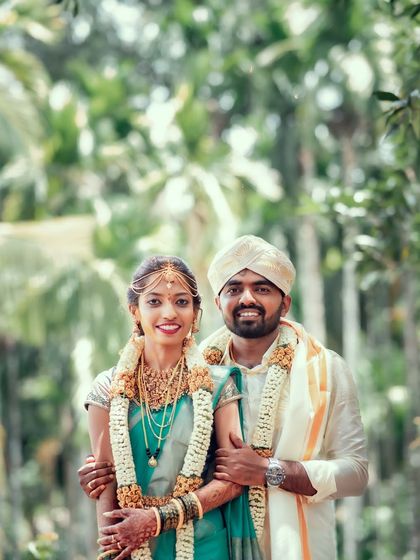 A classic portrait of a smiling South Indian couple, posing together against a backdrop of lush greenery.