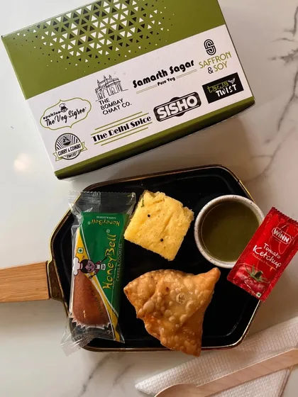 A duplicate of our classic snack box, this image again shows the popular combination of a samosa, dhokla, and a sweet treat. The consistent quality and presentation are key to our service.