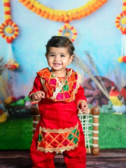 This toddler is all smiles and ready to celebrate in a bright red traditional outfit. The festive backdrop with marigold garlands completes the celebratory look.