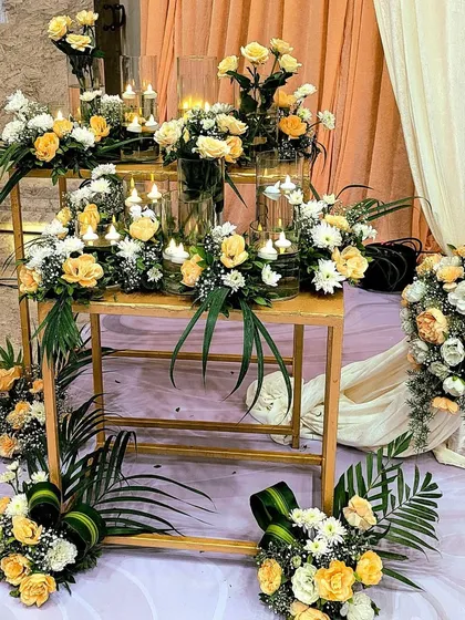 A modern and creative floral installation, where arrangements of yellow and white roses are placed on a multi-level golden stand, surrounded by candles.