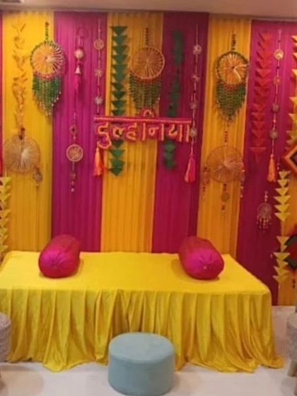 A vibrant backdrop for a "Dulhaniya" or bridal shower event. The decor uses yellow and pink drapes, dreamcatchers, and low seating with poufs for a fun and festive vibe.