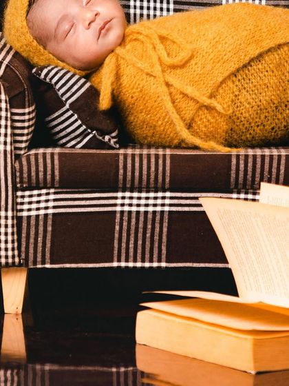 A detailed close-up from our "Advait" setup, focusing on the sleeping baby in a yellow knit outfit on the plaid sofa. The open book in the foreground adds to the studious and charming theme.