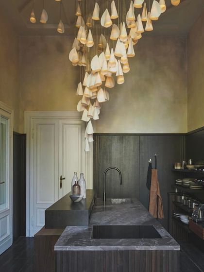 A cascading installation of the Bocci 21 series, made of delicate porcelain, flows down into a modern kitchen. The organic, draped forms contrast beautifully with the clean lines of the dark cabinetry and marble countertops.