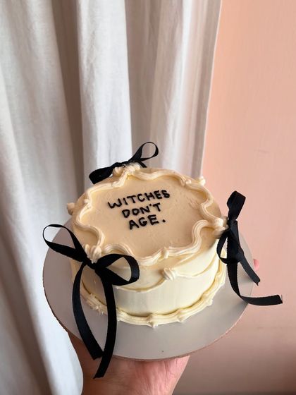 A fun and witchy bento cake with the phrase "Witches don't age." The vintage piping and black bows give it a gothic coquette feel.