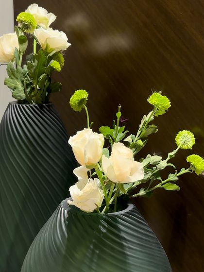 A pair of modern, matte green vases with a spiral texture. The simple arrangements of white ranunculus and green button mums create a sophisticated and understated floral moment.