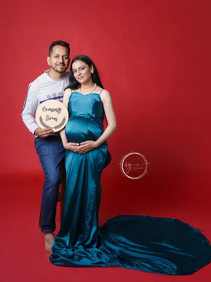 "Coming Soon". This couple uses a sweet prop to announce their little one's impending arrival. The vibrant red background makes the teal gown pop, creating a visually striking maternity announcement.