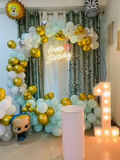 A first birthday decoration for a home, featuring a balloon arch in mint green, white, and gold. The setup includes a light-up marquee number and a Boss Baby foil balloon.