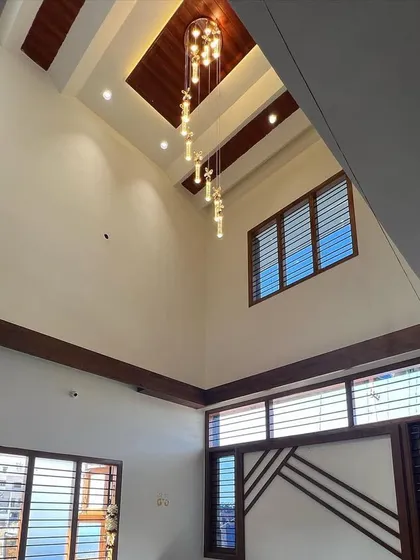 A view of a double-height living space before the final furnishings. The image highlights the architectural details, including the wooden ceiling accents, large windows for natural light, and a beautiful hanging chandelier that serves as a focal point.