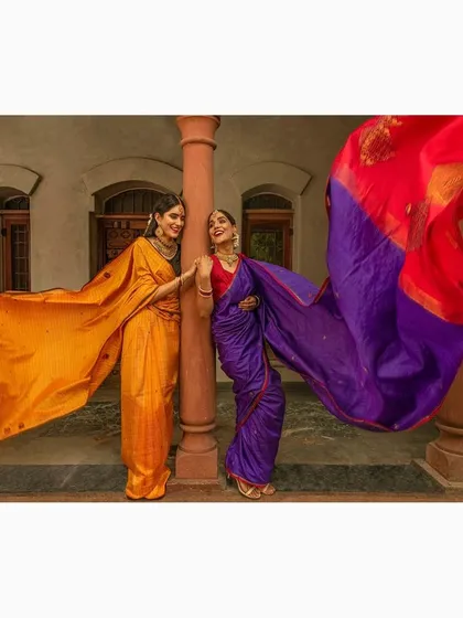A fashion shoot featuring two models in vibrant silk sarees against an architectural backdrop. The wind catching the fabric creates a dramatic, flowing effect that showcases the beauty of the ethnic garments.