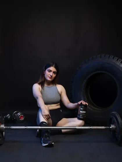 A relaxed post-workout style shot, with the model seated on the floor next to a barbell and the supplement. This image conveys a sense of accomplishment and recovery, targeting customers looking for post-workout nutrition solutions.