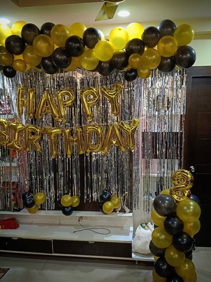 A black and gold birthday decoration for a home, featuring a balloon garland and foil letters over a TV unit.