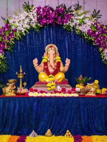 A royal blue sequin backdrop is framed by an arch of purple and white orchids. The floor is decorated with a rangoli of marigold and other flowers, creating a rich and colorful contrast.
