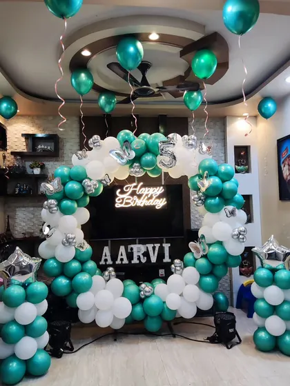 A circular balloon arch in teal and white for a personalized birthday celebration. The setup includes a 'Happy Birthday' neon sign, the birthday girl's name in large letters, and balloon pillars with star toppers.