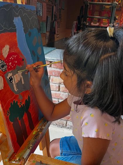 A young girl carefully applies paint to her canvas, building up the layers of color in her vibrant landscape, inspired by the work of David Hockney.