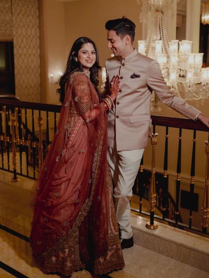 An elegant portrait of the couple on a staircase. The bride's deep red embroidered lehenga and the groom's tailored blush pink Jodhpuri suit create a sophisticated and romantic look for their sangeet or reception.