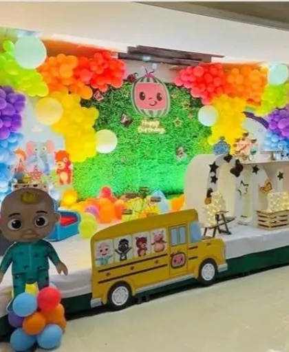 A large-scale Cocomelon stage decoration. This vibrant setup includes a green hedge backdrop, a rainbow of balloons, and multiple character cutouts, including the iconic yellow school bus.