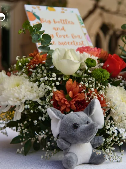 A centerpiece for a 'Little Cutie' themed shower, featuring a small stuffed elephant and a custom card amidst a bouquet of orange and white flowers.