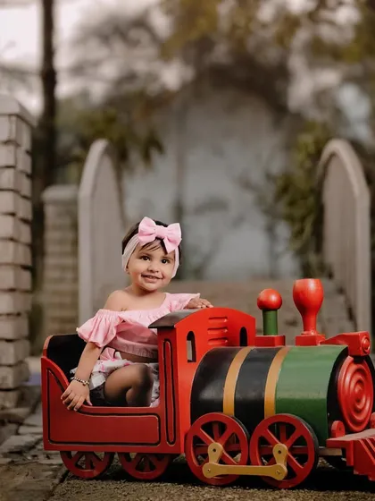 A stylish little girl enjoying a ride on a toy train. The bridge in the background adds a lovely scenic element to this outdoor portrait.