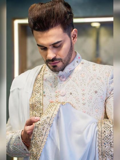 A moment of admiration for the craft. Ayush Khatri inspects the detailed embroidery on the stole of his custom white sherwani, a piece designed for ultimate sophistication.