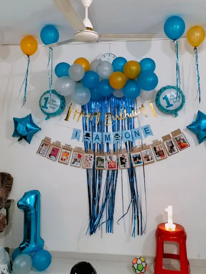 A simple and effective first birthday decoration for a home. It includes a foil curtain backdrop, a photo banner showing the baby's first year, and blue-themed balloons.