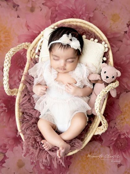 A vision in white lace. This newborn girl, dressed in a delicate outfit, sleeps in a basket against a backdrop of pink flowers.